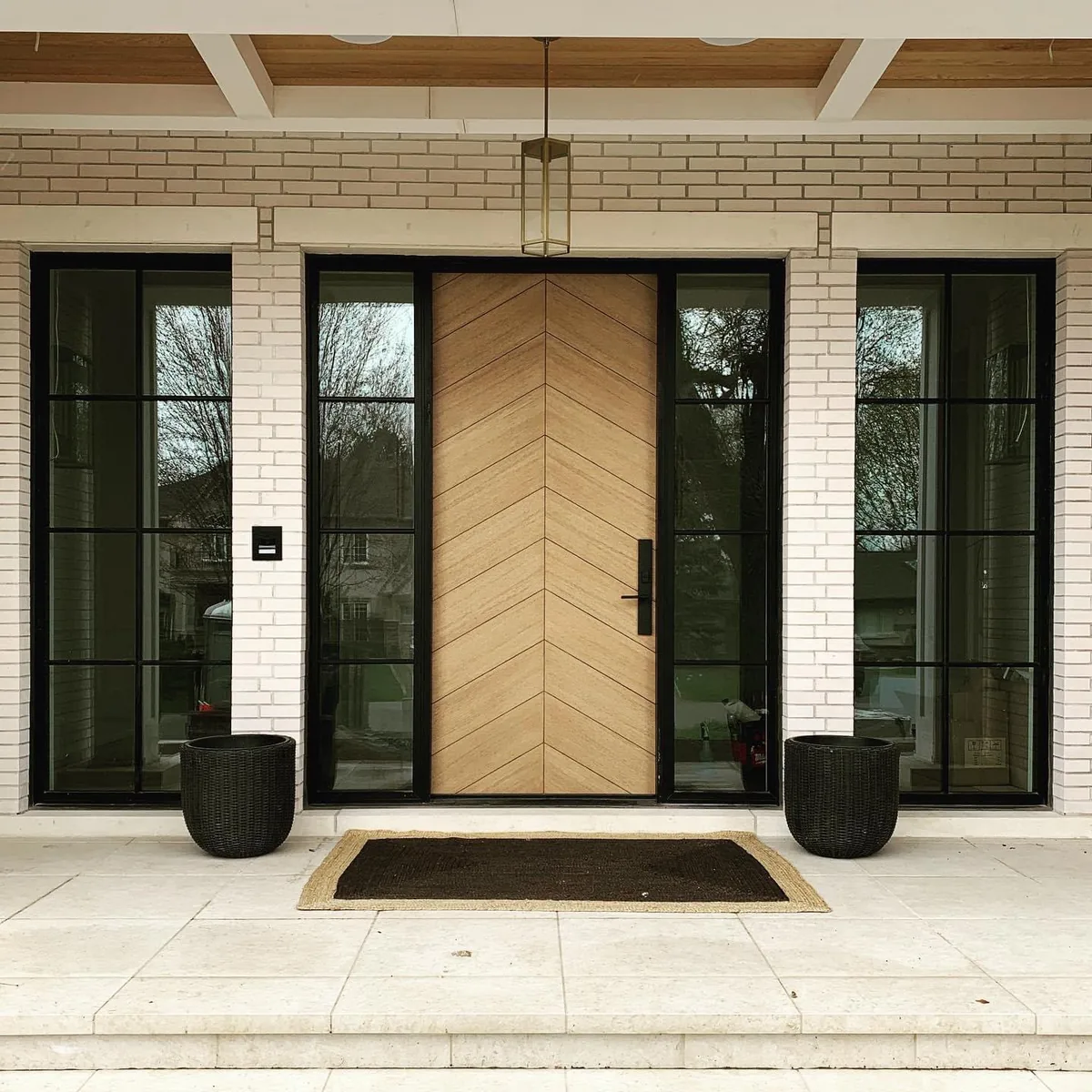 Custom herringbone wood entry door with black steel-grid glass sidelites on a modern white-brick home, custom door manufacturing in Toronto