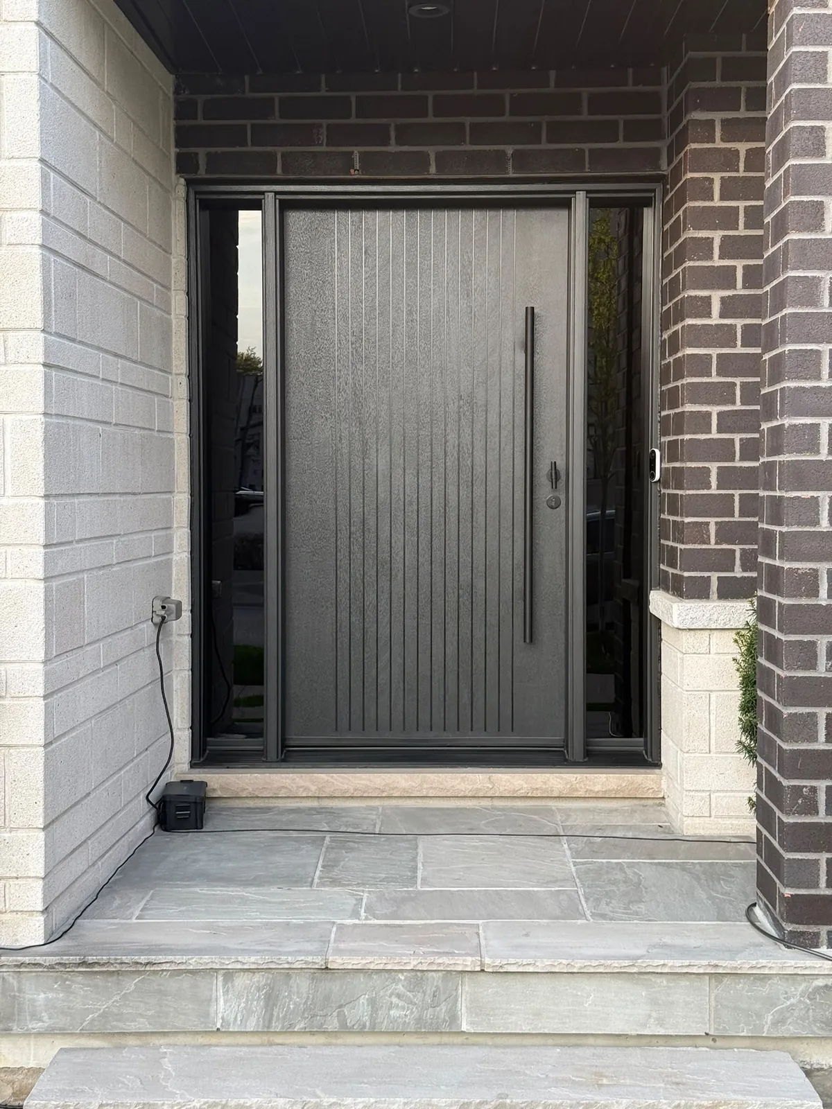 Modern grey vertical-groove entry door with a full-height bar handle and glass sidelite, custom modern doors in Toronto