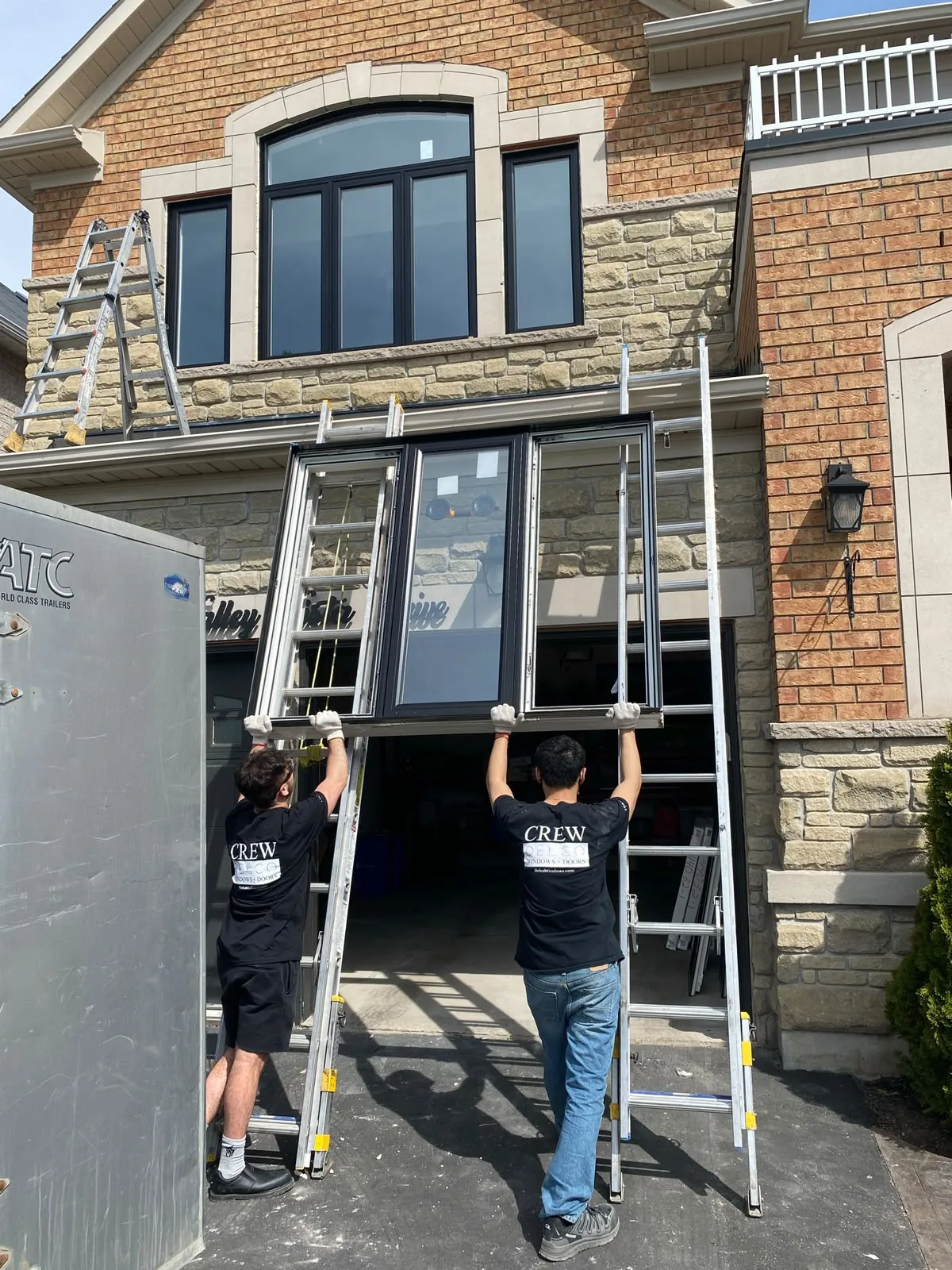 Two Delco crew members carrying a large new window unit up on ladders at a brick and stone home, window installation in Vaughan